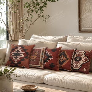 May include: Five square throw pillows arranged on a light-colored sofa. Each pillow features a distinct geometric pattern in shades of rust, terracotta, black, and cream. The designs include diamond motifs, stepped borders, and stylized animal figures.