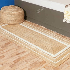 Könnte beinhalten: Ein Naturfaser-Teppich mit weißem Rand. Der Teppich ist aus Jute und hat eine natürliche, erdige Farbe. Es ist ein rechteckiger Teppich mit einer langen, schmalen Form. Der Teppich liegt auf einem Holzboden.