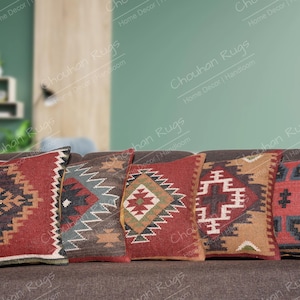 Puede incluir: Cinco cojines decorativos con patrones geométricos en tonos rojos, marrones, negros y blancos. Los cojines están dispuestos en un sofá marrón.