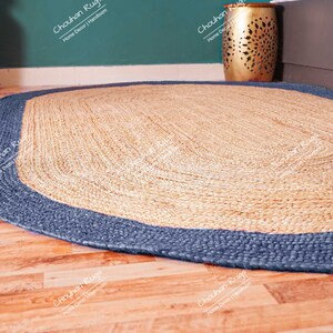 K&ouml;nnte beinhalten: Ein ovaler Teppich aus Naturfasern mit einem blauen Rand. Der Teppich ist aus gewebtem Jute gefertigt und hat eine nat&uuml;rliche, erdige Farbe.