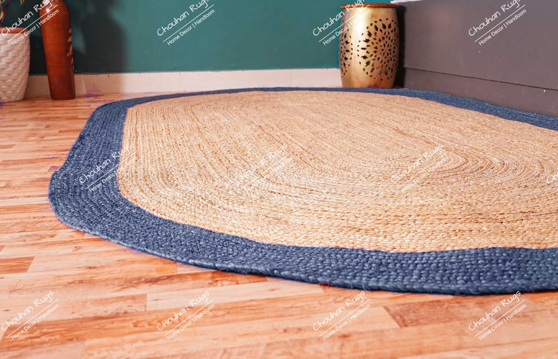 K&ouml;nnte beinhalten: Ein ovaler Teppich aus Naturfasern mit einem blauen Rand. Der Teppich ist aus gewebtem Jute gefertigt und hat eine nat&uuml;rliche, erdige Farbe.