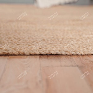 May include: A close-up of a natural fiber rug with a woven texture. The rug is light brown and has a braided edge. The rug is lying on a wooden floor.