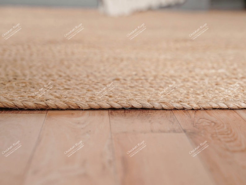 May include: A close-up of a natural fiber rug with a woven texture. The rug is light brown and has a braided edge. The rug is lying on a wooden floor.