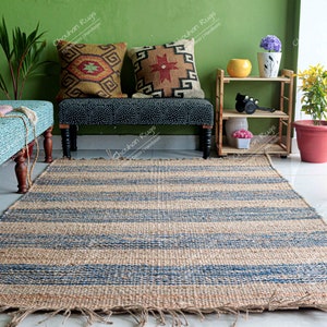 K&ouml;nnte beinhalten: Ein blauer und naturfarbener Jute-Streifen-Teppich mit ausgefransten Kanten. Der Teppich liegt vor einer hellblauen Bank mit zwei gemusterten Kissen. Die Bank steht vor einer gr&uuml;nen Wand mit einem kleinen Holzregal mit Pflanzen und einer gelben Schale.
