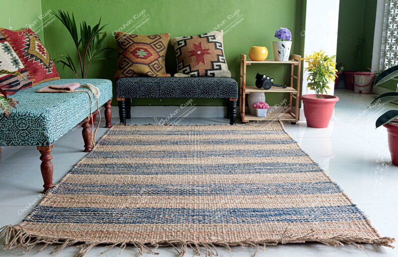 K&ouml;nnte beinhalten: Ein blauer und naturfarbener Jute-Streifen-Teppich mit ausgefransten Kanten. Der Teppich liegt vor einer hellblauen Bank mit zwei gemusterten Kissen. Die Bank steht vor einer gr&uuml;nen Wand mit einem kleinen Holzregal mit Pflanzen und einer gelben Schale.