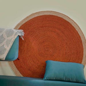 Könnte beinhalten: Ein großer runder Teppich mit einer leuchtend rostorangen Mitte, umgeben von einem naturfarbenen Juterand. Der Teppich befindet sich hinter einem blaugrünen Sofa mit einem passenden rechteckigen Kissen und einer weißen Decke mit grauem Blattmuster.