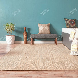 May include: A light brown jute rug with a woven texture, placed on a wooden floor in front of a teal wall. A wooden bench with a grey cushion and a throw pillow sits in the middle of the rug. A white planter with a green plant sits on the left side of the rug. A dark brown bed with a white sheet and a cream throw blanket with a palm tree pattern sits on the right side of the rug.