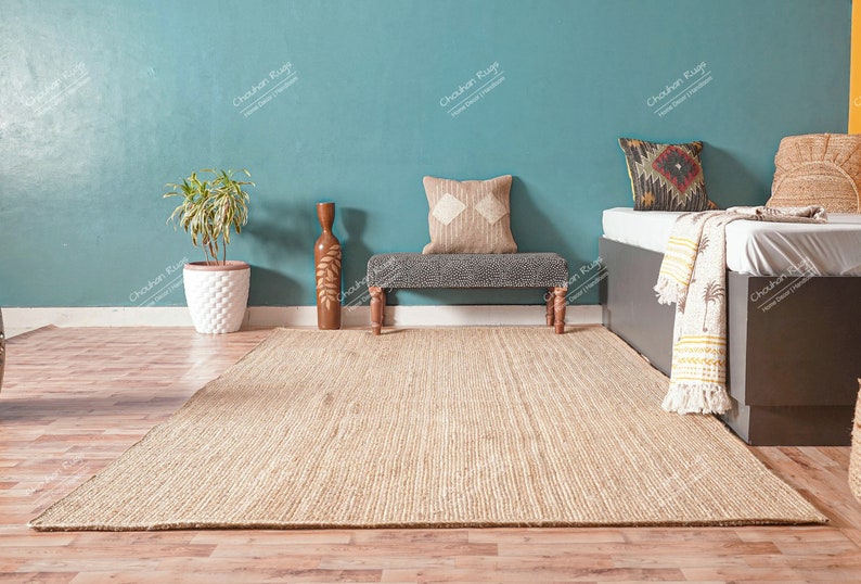 May include: A light brown jute rug with a woven texture, placed on a wooden floor in front of a teal wall. A wooden bench with a grey cushion and a throw pillow sits in the middle of the rug. A white planter with a green plant sits on the left side of the rug. A dark brown bed with a white sheet and a cream throw blanket with a palm tree pattern sits on the right side of the rug.