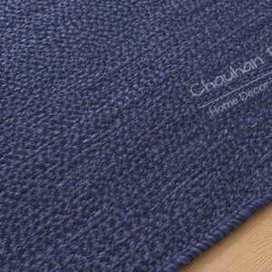 Può includere: Un tappeto intrecciato blu scuro con una superficie testurizzata e un bordo rifinito. Il testo "Chauhan Rugs Home Decor | Handloom" è stampato in bianco sul lato superiore destro del tappeto, posizionato sopra una superficie in legno chiaro.