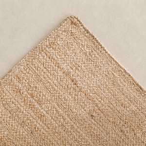 May include: A rectangular area rug featuring a light tan, natural fiber weave with a dense, pebbled texture. The rug has a clean, finished edge with a matching stitched border that defines the corner and maintains a consistent, uniform appearance.