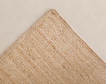 May include: A rectangular area rug featuring a light tan, natural fiber weave with a dense, pebbled texture. The rug has a clean, finished edge with a matching stitched border that defines the corner and maintains a consistent, uniform appearance.
