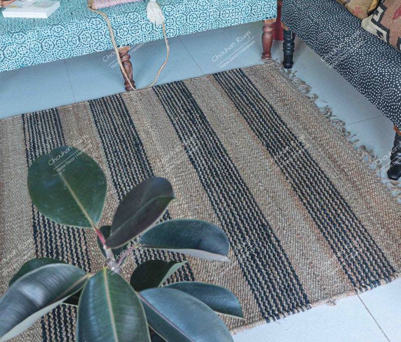 K&ouml;nnte beinhalten: Ein Naturfaser-Teppich mit schwarzen und braunen Streifen. Der Teppich ist aus Jute und hat einen Fransenrand. Eine gr&uuml;ne Pflanze befindet sich im Vordergrund.