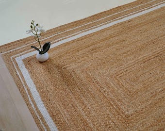 May include: A rectangular area rug woven from natural tan jute fibers, featuring a distinct white border stripe pattern that follows the rectangular shape of the rug. A small white potted orchid with white flowers sits on the rug surface.