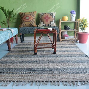 K&ouml;nnte beinhalten: Ein gestreifter Jute-Teppich mit Fransen an den R&auml;ndern in einem Wohnzimmer. Der Teppich liegt vor einer Holzbank mit blau-wei&szlig; gemusterter Polsterung und zwei Kissen mit geometrischen Mustern. Ein kleiner Holztisch mit einer geflochtenen Tischplatte steht auf dem Teppich.