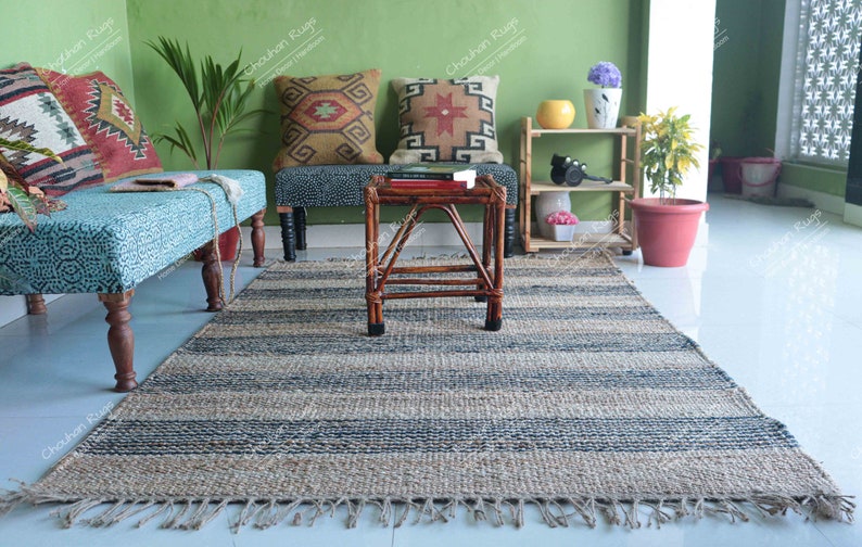 K&ouml;nnte beinhalten: Ein gestreifter Jute-Teppich mit Fransen an den R&auml;ndern in einem Wohnzimmer. Der Teppich liegt vor einer Holzbank mit blau-wei&szlig; gemusterter Polsterung und zwei Kissen mit geometrischen Mustern. Ein kleiner Holztisch mit einer geflochtenen Tischplatte steht auf dem Teppich.