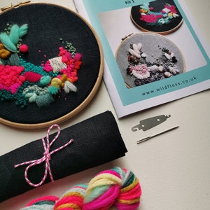 Op de afbeelding: Een borduurpakket met een afgewerkt stuk in een houten hoepel, met levendig roze, teal en geel garen. De kit bevat een zwarte stoffen rol vastgebonden met roze en wit touw, een streng kleurrijk garen en een boekje met de tekst "Wild Floss Embroidery".