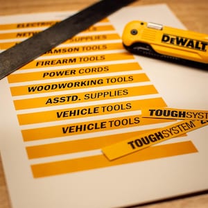 Puede incluir: Una hoja blanca con etiquetas horizontales amarillas con texto negro que incluye Electric, Network Supplies, Samson Tools, Firearm Tools, Power Cords, Woodworking Tools, Asstd. Supplies, Vehicle Tools y Toughsystem, junto a un cúter amarillo DeWalt.