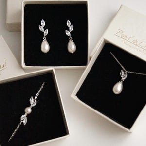 Può includere: Un assortimento di set di gioielli da sposa, tra cui orecchini, collana e bracciale, presentati in scatole color crema con la scritta "Pearl & Crystal Wedding Accessories". Ogni pezzo presenta una perla a goccia e dettagli in cristallo.