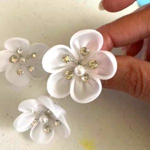May include: Three white fabric flower embellishments with pearl and rhinestone accents.