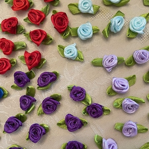 May include: A collection of small, fabric rose embellishments in various colors. The roses are red, light blue, purple, and lavender, each with a green leaf backing. These decorative flowers are ideal for crafts.