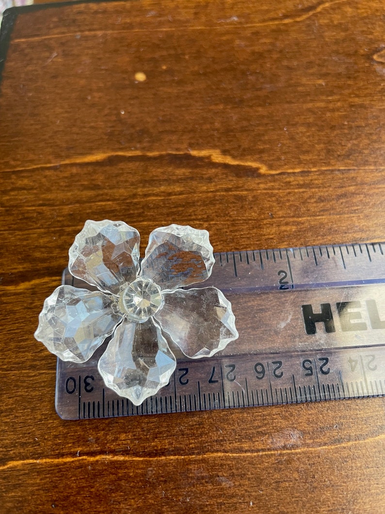 May include: A clear crystal flower with five petals and a small crystal centre. The flower is approximately 5 cm wide and is resting on a ruler.