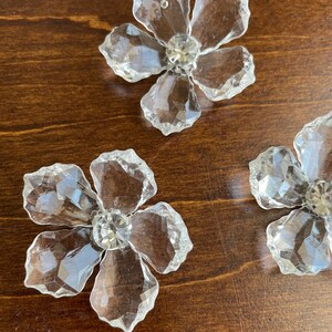 May include: Three clear crystal flower-shaped embellishments with a faceted design and a clear rhinestone in the centre of each flower.