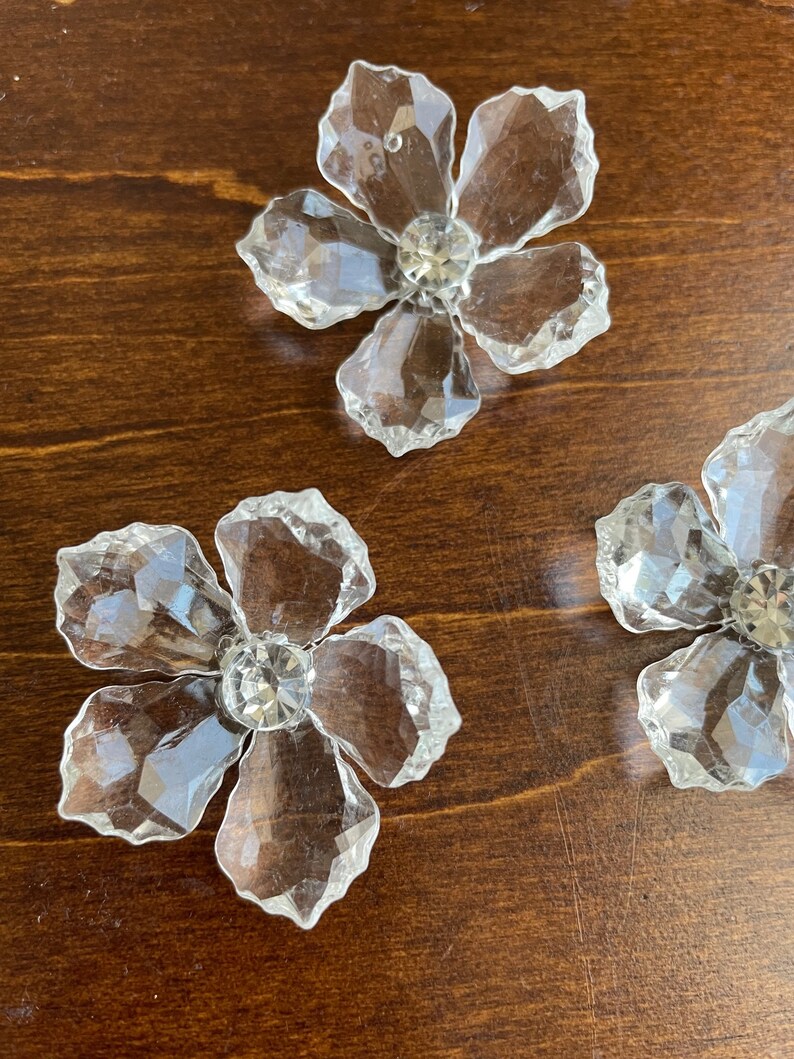 May include: Three clear crystal flower-shaped embellishments with a faceted design and a clear rhinestone in the centre of each flower.
