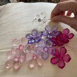 May include: A collection of crystal flower embellishments in various colours, including pink, purple, and clear. The flowers have a faceted design and are arranged on a white fabric surface.