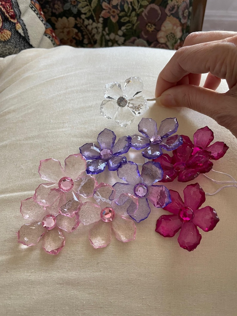 May include: A collection of crystal flower embellishments in various colours, including pink, purple, and clear. The flowers have a faceted design and are arranged on a white fabric surface.