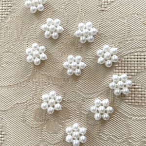 May include: Ten white pearl flower embellishments on a beige fabric background.
