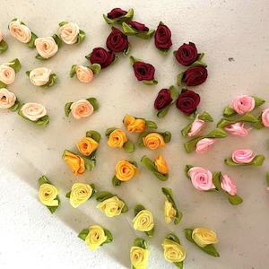 May include: A collection of small, fabric rose embellishments in various colors. The roses are in shades of pink, yellow, and burgundy, each with green leaf accents. These decorative flowers are ideal for crafting and embellishing projects.