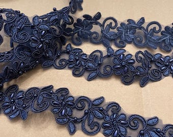 May include: Navy blue floral lace trim with beaded accents. The trim features a delicate floral pattern with intricate details and small, round beads. The lace is arranged in a flowing, decorative manner, suitable for various craft projects.