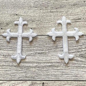 May include: Two white embroidered crosses with a decorative design. The crosses are on a light wood background.