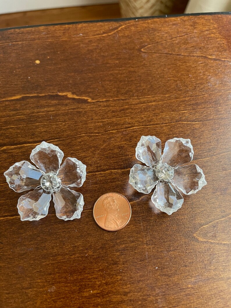 May include: Two clear crystal flower-shaped embellishments with a faceted design. The embellishments are approximately the size of a UK penny.