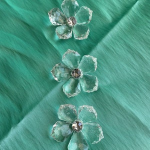 May include: Three clear glass flower-shaped ornaments with a crystal centre on a teal background.