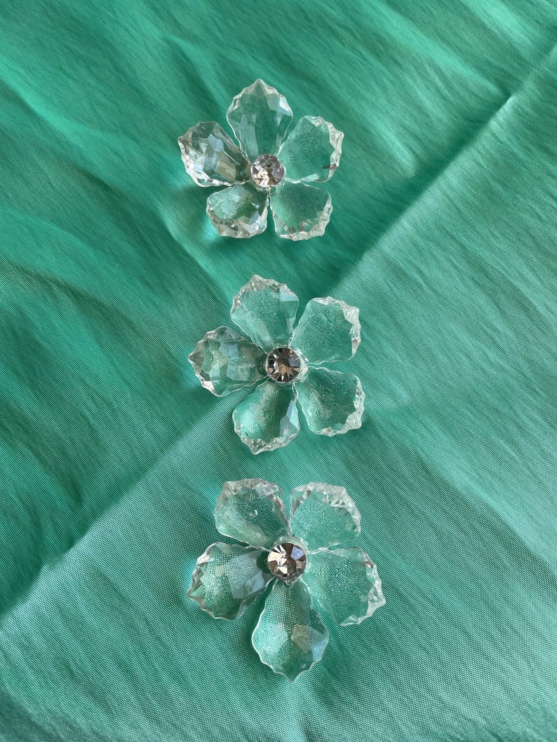 May include: Three clear glass flower-shaped ornaments with a crystal centre on a teal background.