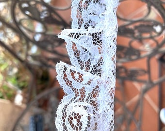 May include: Close-up of white lace trim featuring a delicate floral design. The lace has a scalloped edge and is made of a fine, open weave material. The trim is set against a blurred background.