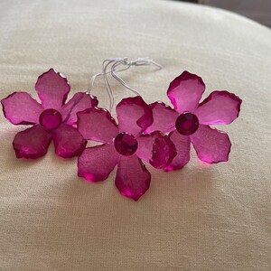 May include: Four pink crystal flower ornaments with clear rhinestone centres. The flowers are attached to white strings.