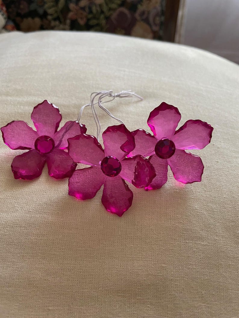 May include: Four pink crystal flower ornaments with clear rhinestone centres. The flowers are attached to white strings.