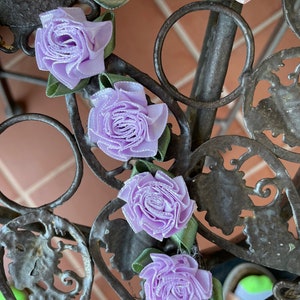 May include: Close-up of a decorative metal object with ribbon roses. The roses are in shades of pink and lavender, with green ribbon leaves. The metal has an aged, rustic appearance.