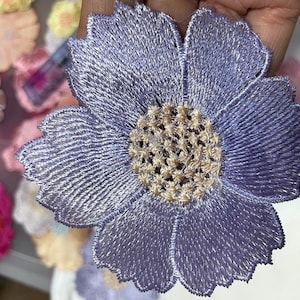 May include: A purple embroidered flower applique with a detailed center. The flower has a delicate, lacy texture and is perfect for adding a touch of elegance to crafts and projects.