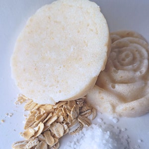 May include: Two white round soap bars with a swirl design, one on top of the other, with a pile of oats and white salt below.