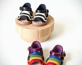 May include: Two pairs of miniature leather sandals. One pair is black and white striped with a black strap. The other pair has a rainbow colored strap and a purple strap.
