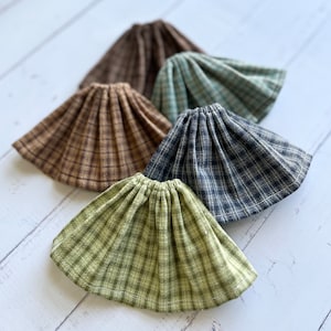 Può includere: Cinque gonne di bambola in tessuto a quadri di vari colori, tra cui marrone, verde, blu e grigio. Le gonne sono disposte in cerchio, con la gonna marrone al centro.