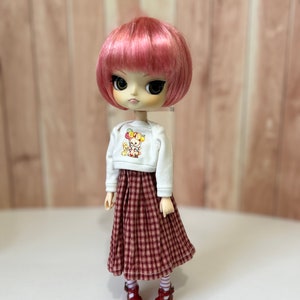 May include: A doll with pink hair wearing a white shirt with a cartoon animal design and a red and white gingham skirt. The doll is wearing red shoes with white socks.