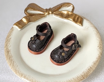 May include: A pair of miniature brown leather Mary Jane shoes with flower accents and buckle closures. The shoes have a textured appearance and are set on a cream-colored plate with a gold bow design.
