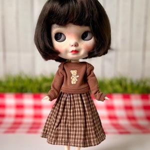 May include: A brown and white checked skirt with a brown jumper featuring a small teddy bear graphic. The doll is wearing brown shoes.