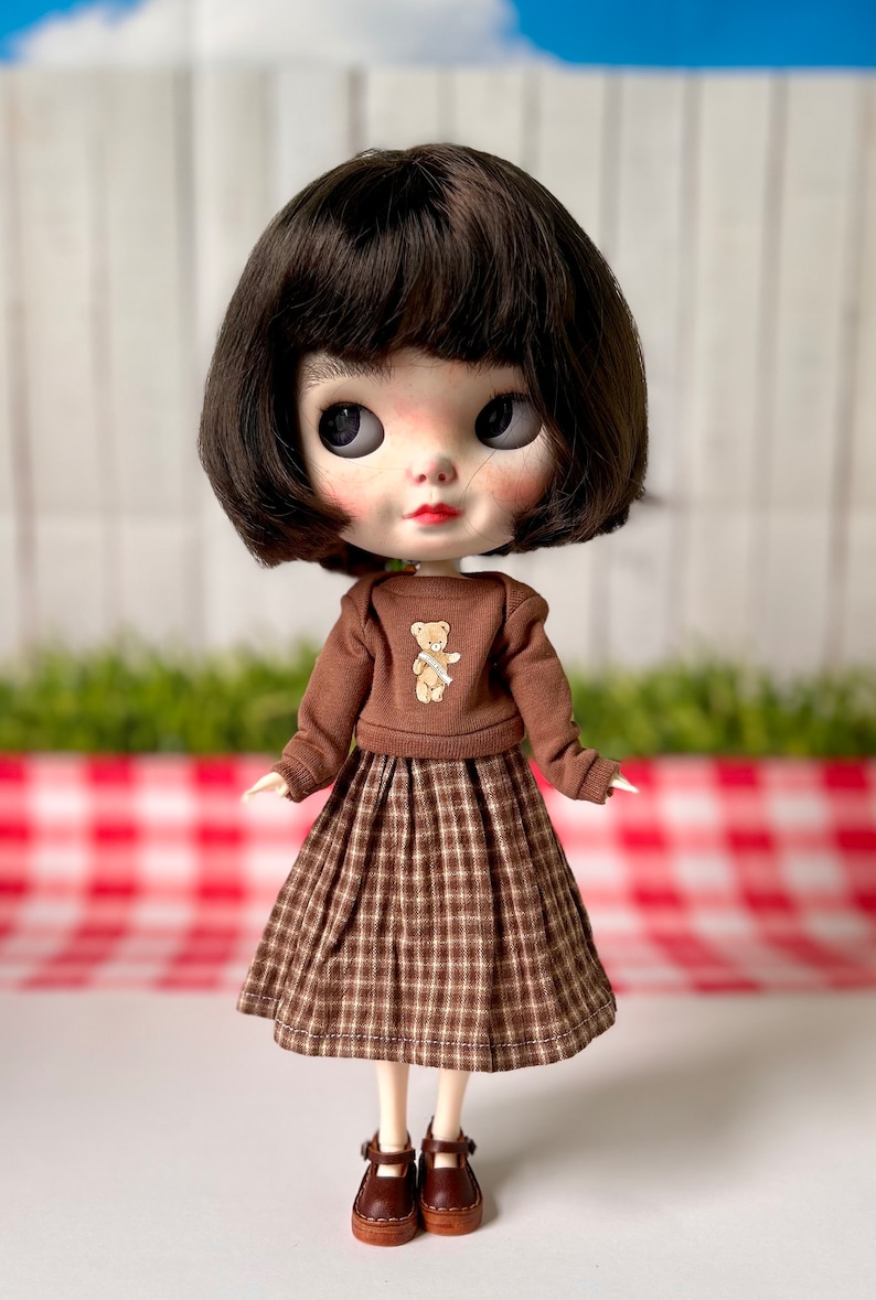 May include: A brown and white checked skirt with a brown jumper featuring a small teddy bear graphic. The doll is wearing brown shoes.