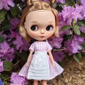 May include: A doll with blonde hair in pigtails wearing a pink and white gingham dress with a white apron. The doll is standing in front of a bush of pink flowers.