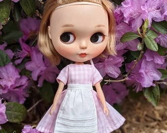 May include: A doll with blonde hair in pigtails wearing a pink and white gingham dress with a white apron. The doll is standing in front of a bush of pink flowers.